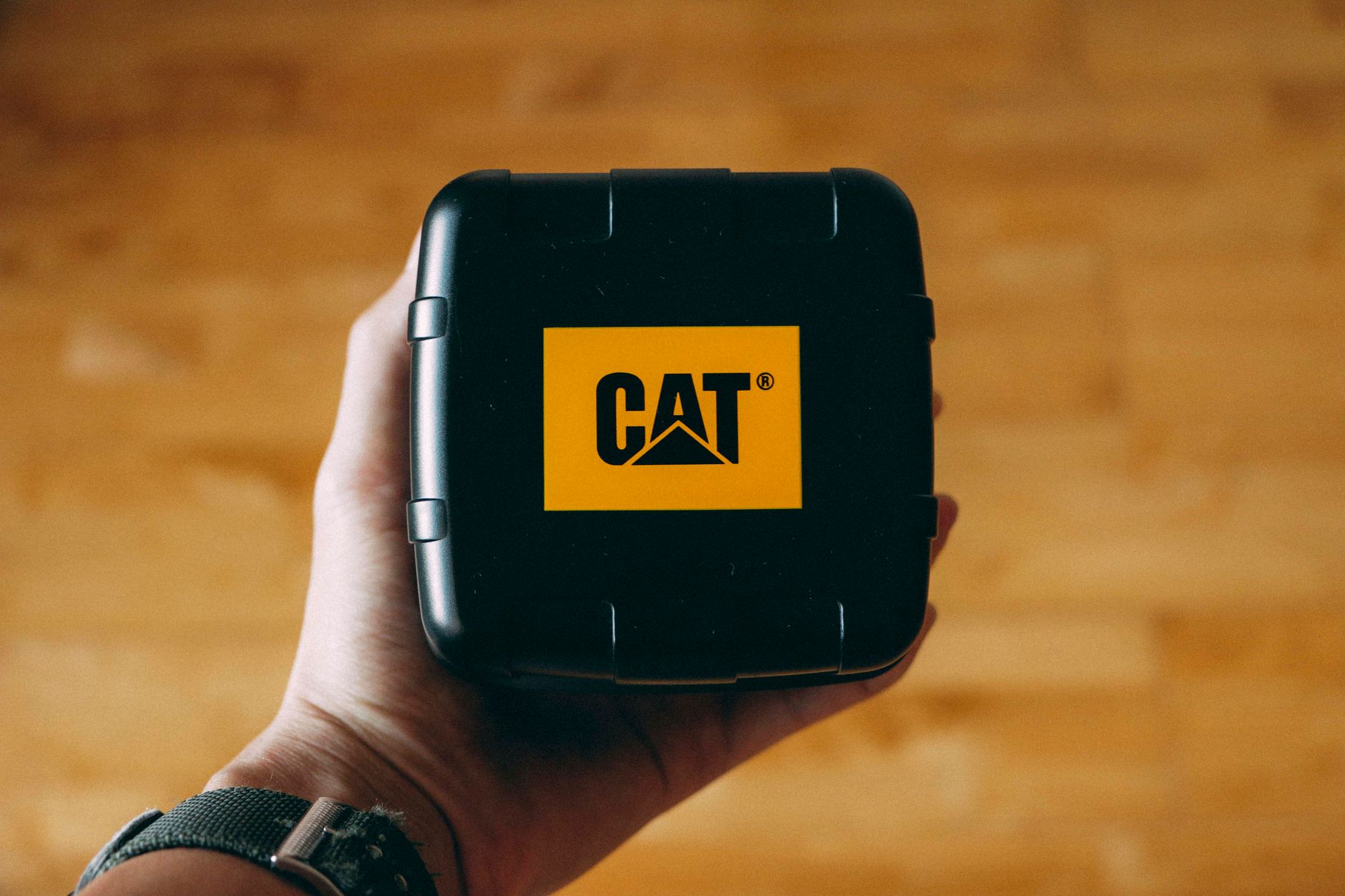 Close-up of a hand holding a black box with CAT branding over a wooden floor. Perfect for industrial and product themes. — Ca