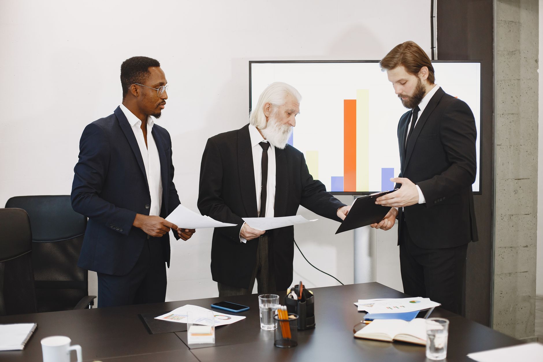 Three businessmen discussing documents in a conference setting with a presentation screen. — Communication Compliance In A Po