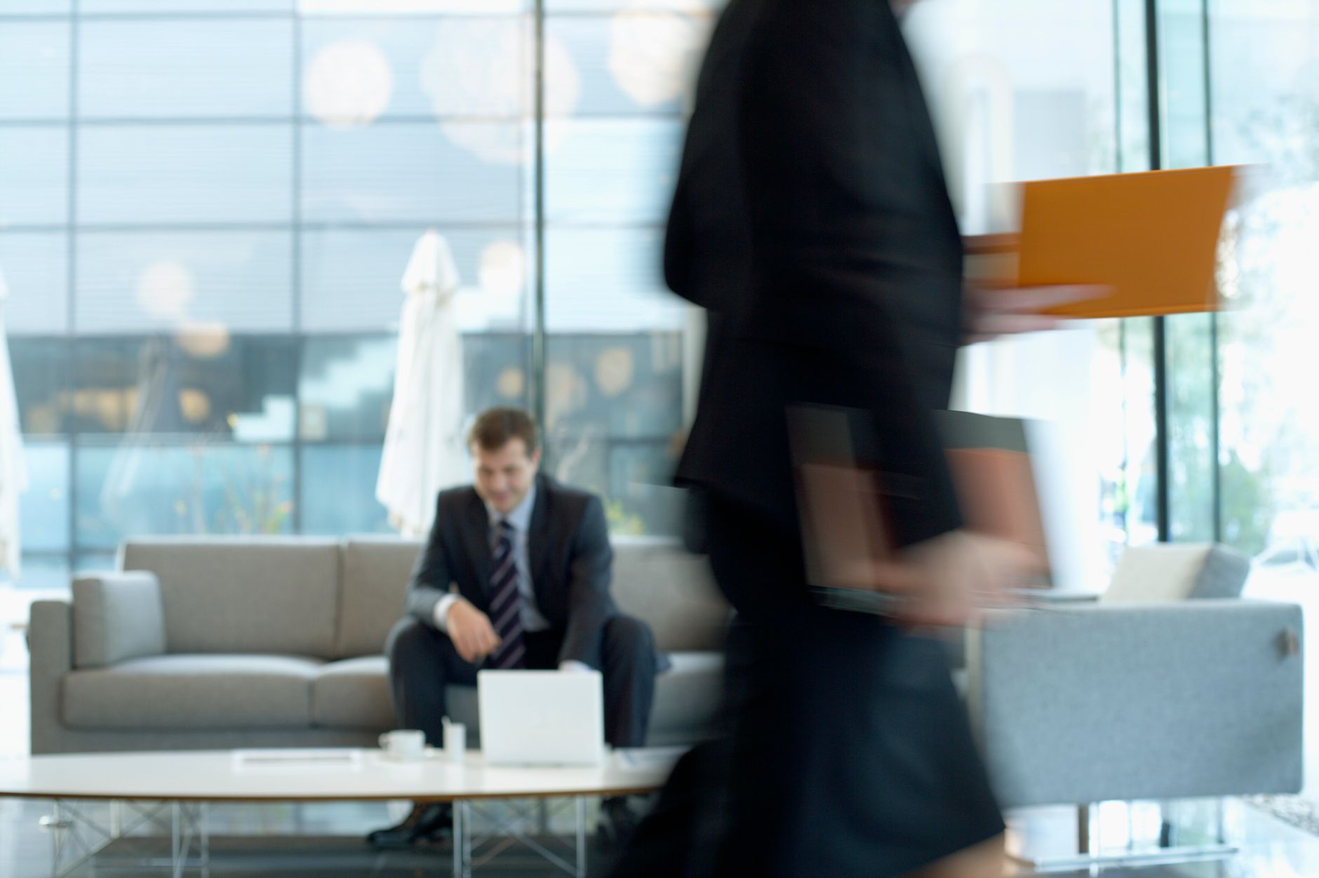 Businessman sitting in modern office lounge using a laptop while colleague walks past, creating dynamic blur. — Factory Audit
