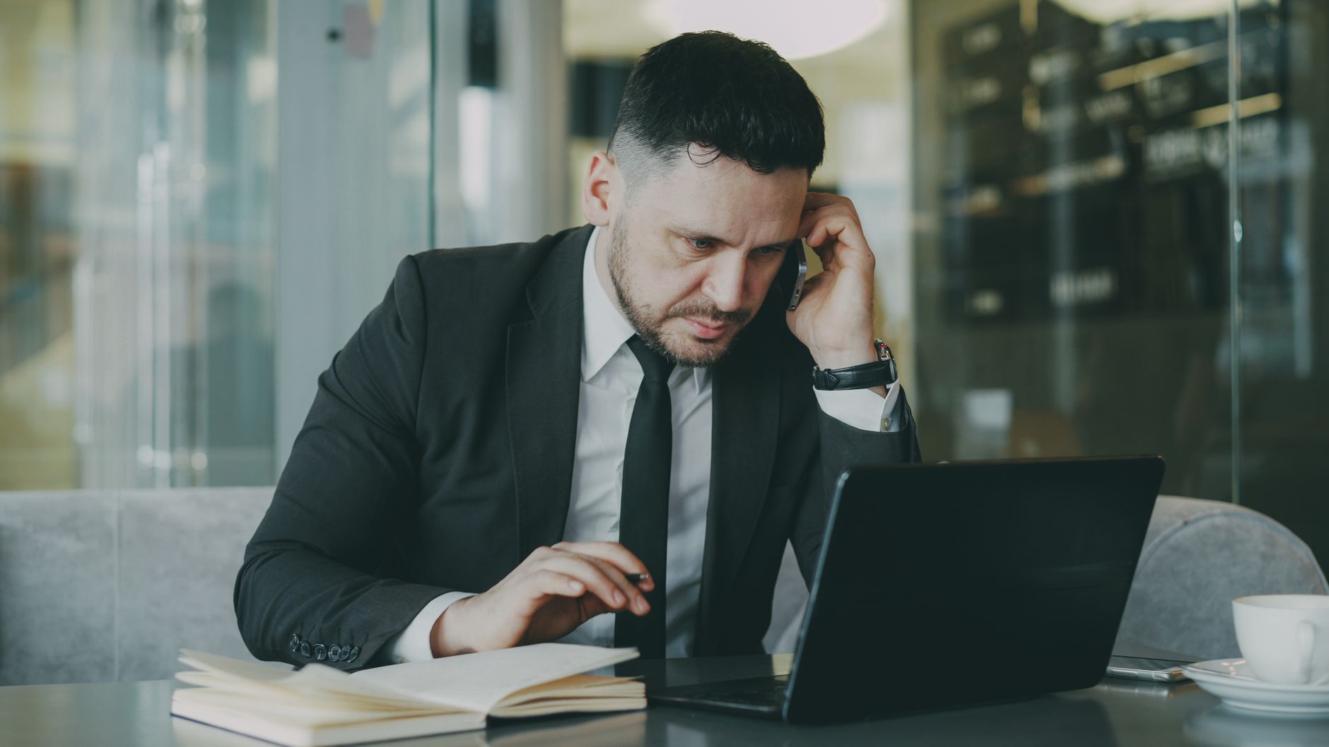 Focused businessman in suit multitasking on phone and laptop in modern office. — How does a corner desk saves space