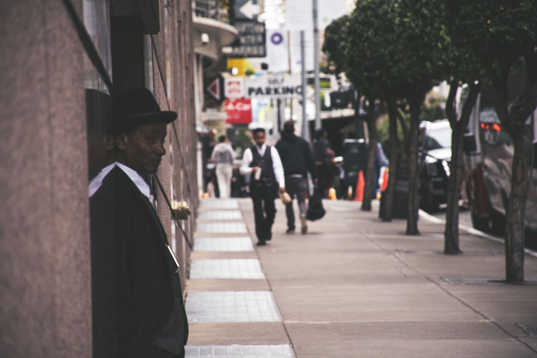 A man in a suit and hat leans against a city building, observing pedestrians. — Is your office buzzing with a strong morale a