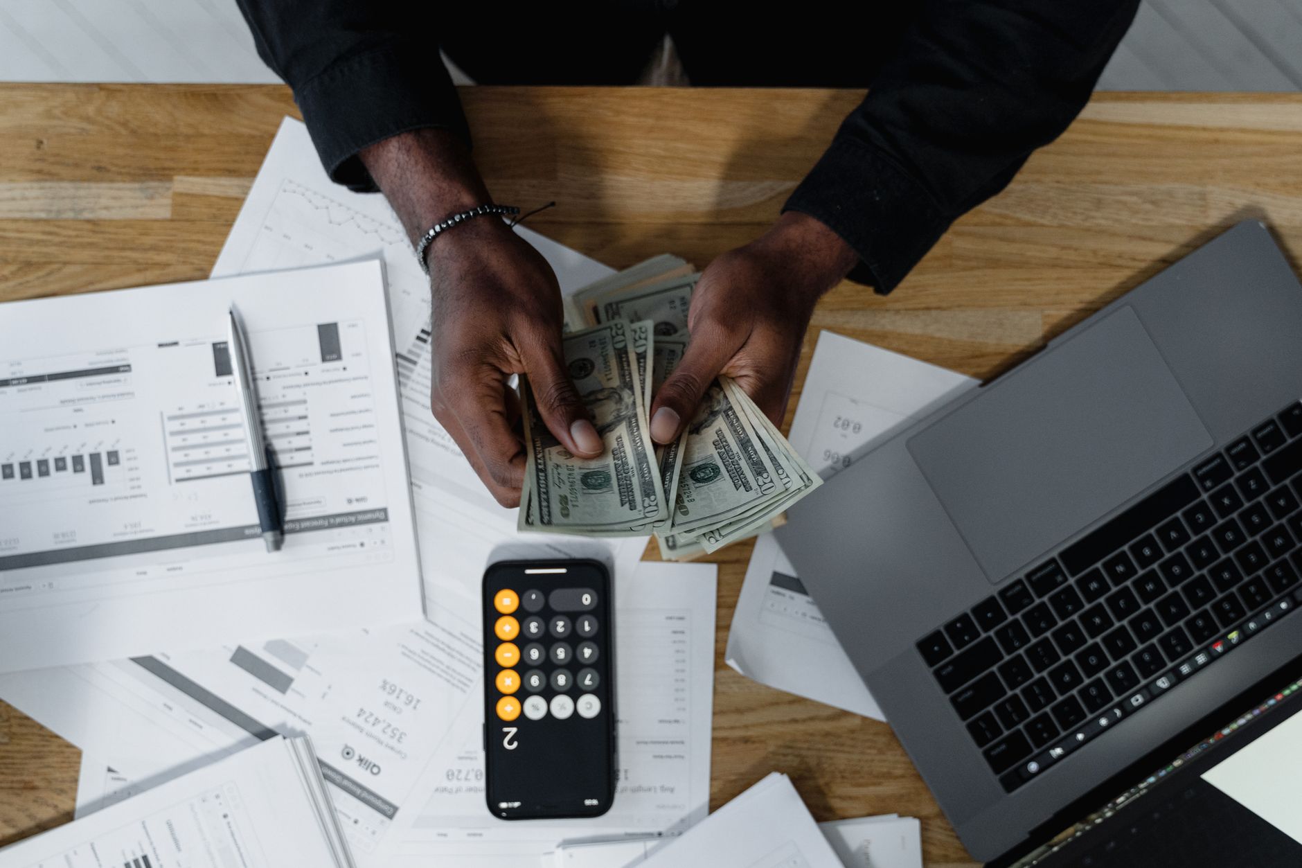 Person counting US dollars, using a calculator and laptop, with financial documents on a wooden desk. — Need a Home Loan? Cho