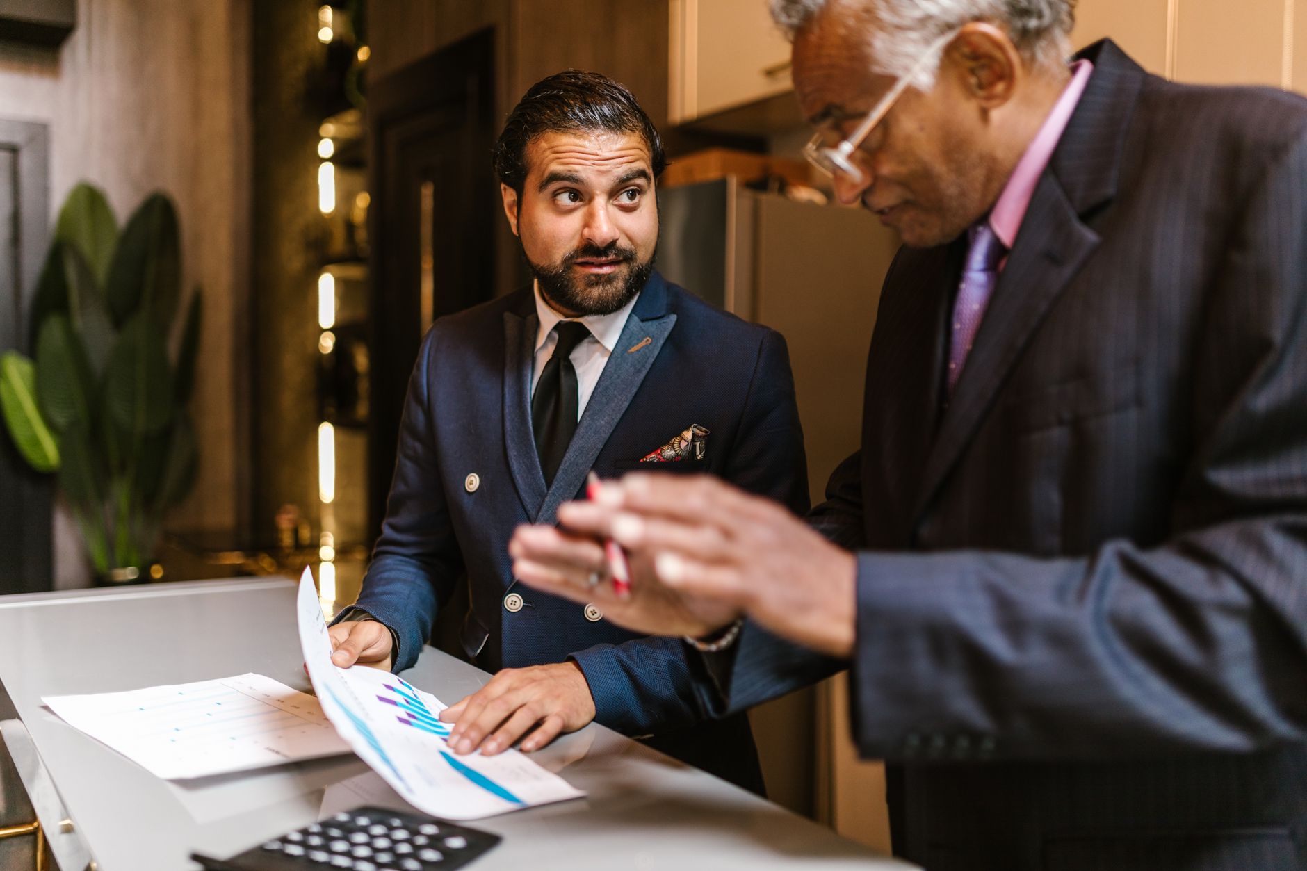 Two men in suits discuss financial documents with graphs indoors. — NEW TO EQUITY FUNDS? START WITH ELSS