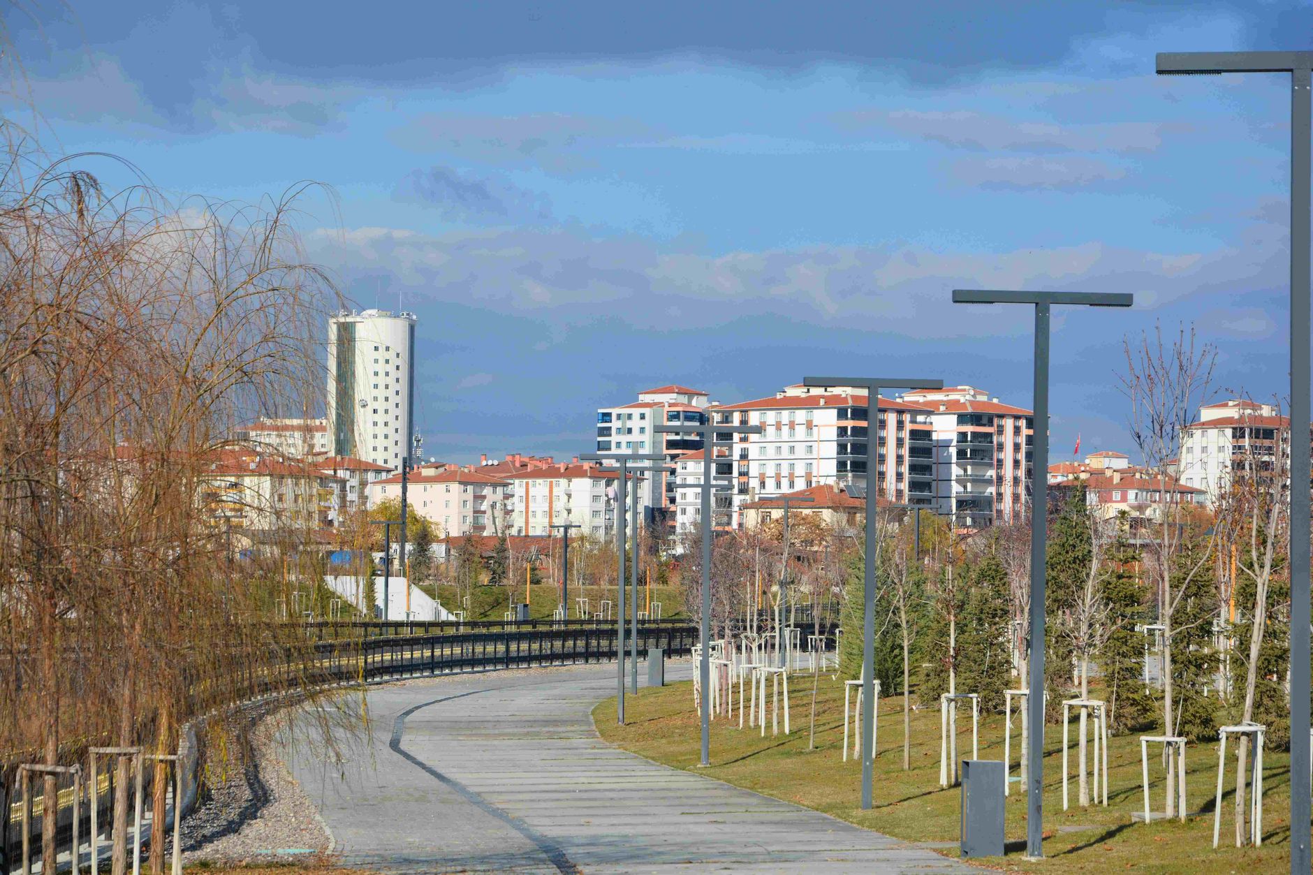 An empty urban park pathway lined with trees, lampposts, and modern residential buildings. — Should you hire an estate planni