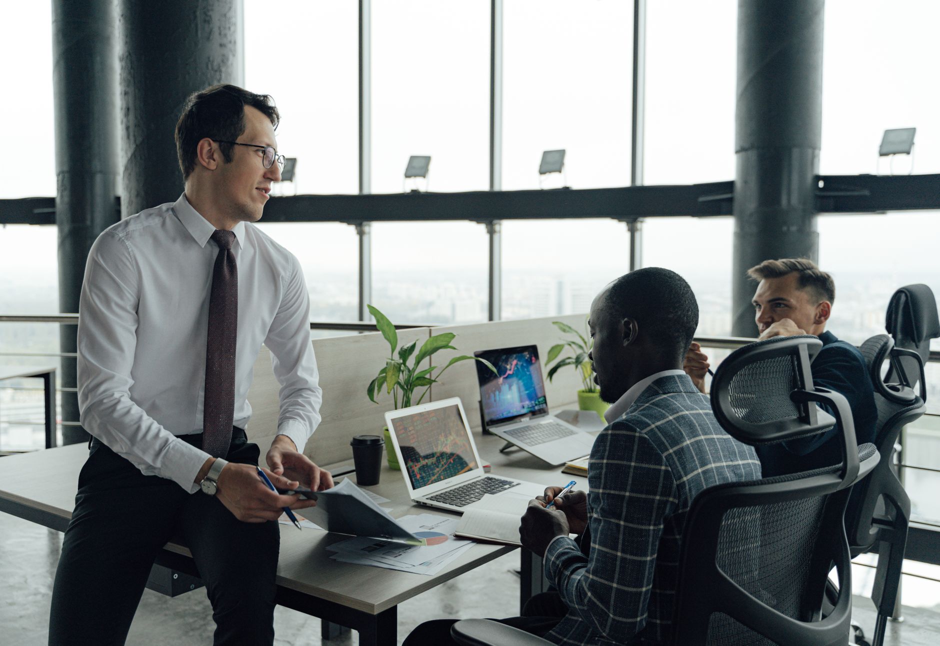 Three men engaged in a business meeting in a modern office setting with laptops and documents. — The effects technological ad
