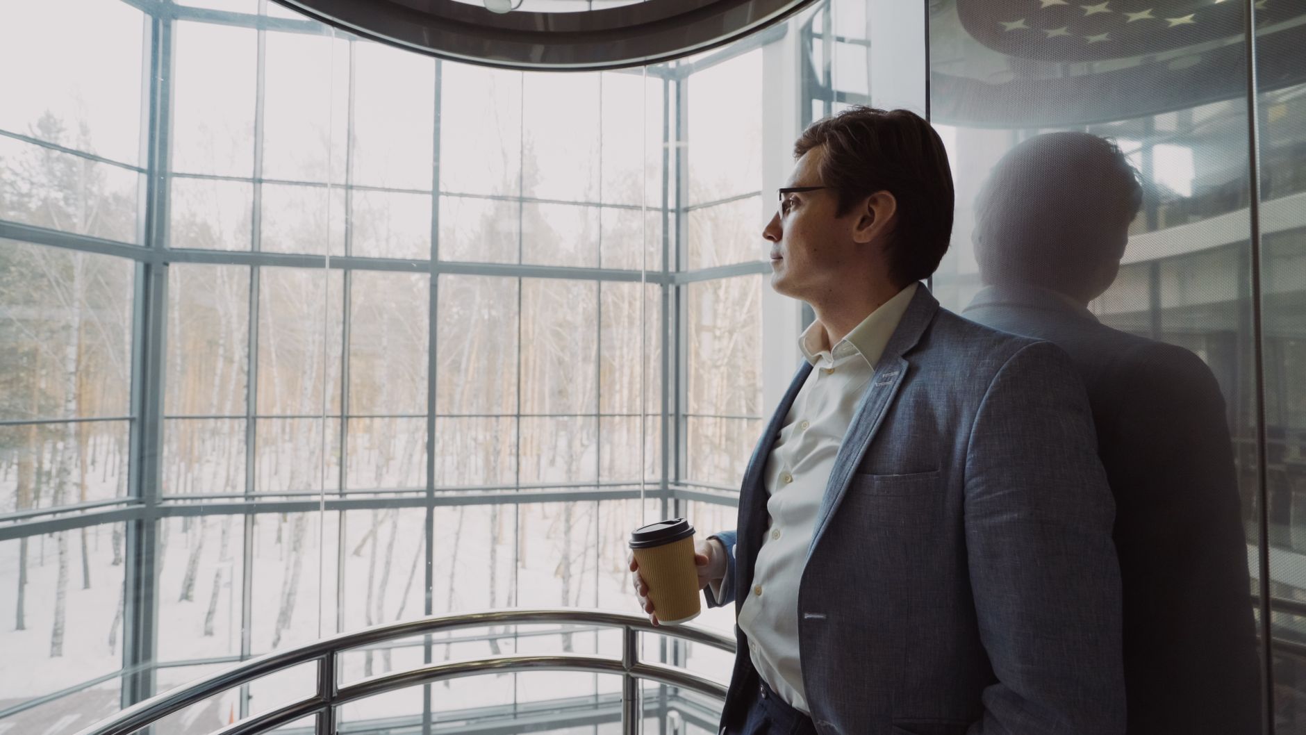Businessman in blue suit with coffee cup looking out a large window, reflecting on a winter landscape. — The Immediate Benefi