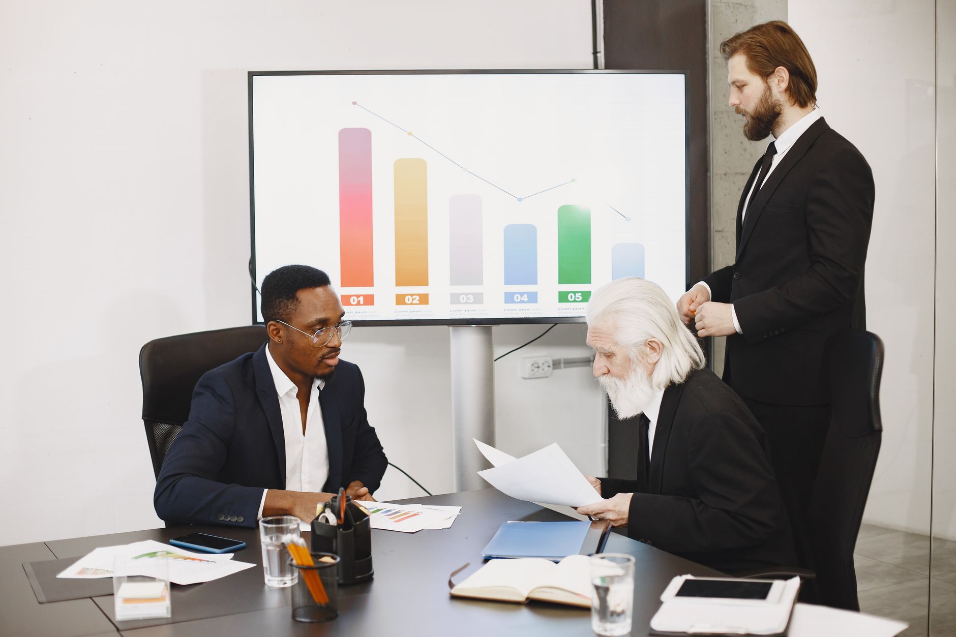 Three businessmen discussing financial data in a modern office setting with charts. — The only business that has benefited fr