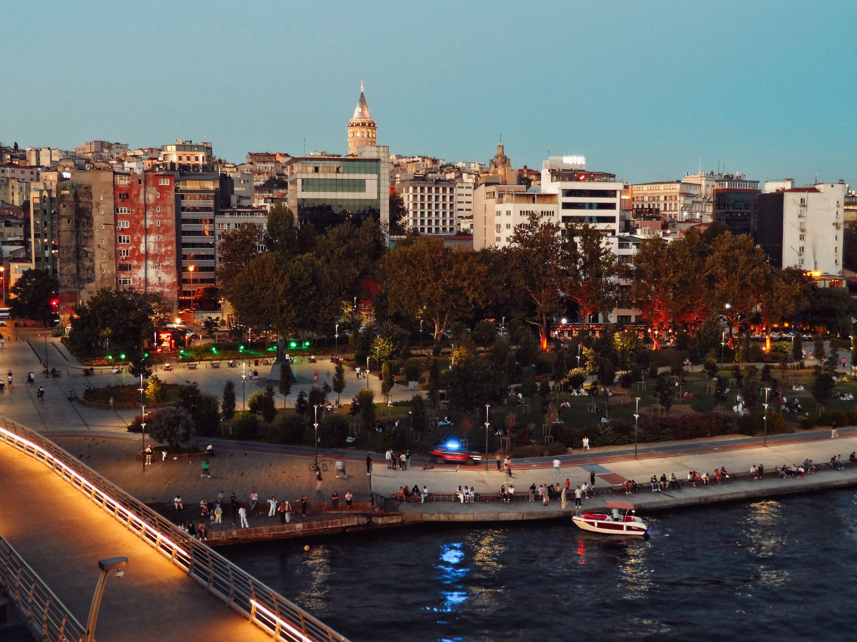 Scenic view of Istanbul's skyline featuring Galata Tower and bustling park at twilight along the waterfront. — Using Frase Re