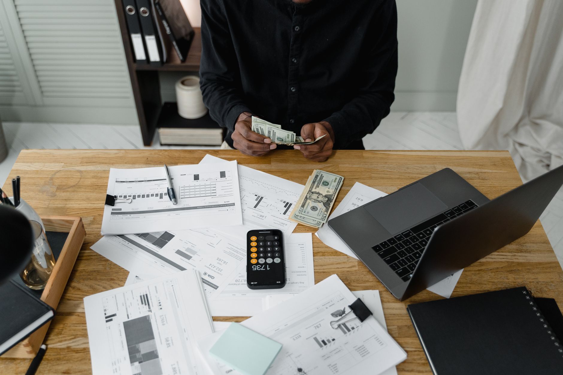 A person counts dollar bills at a desk with a laptop, papers, and calculator. — Very best in Class Finance Functions For Poli