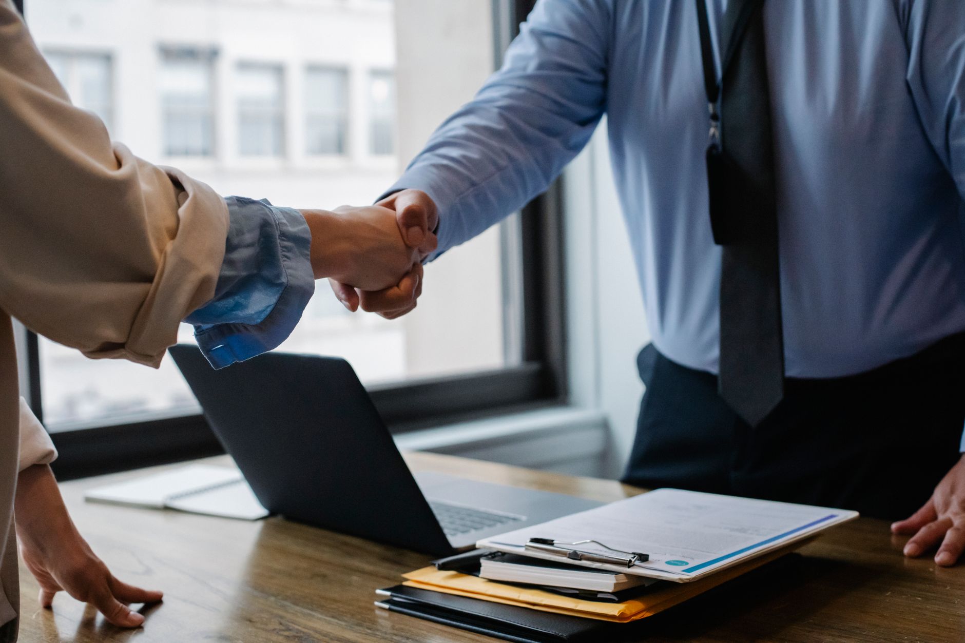 Crop unrecognizable coworkers in formal wear standing at table with laptop and documents while greeting each other before mee